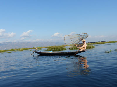 Ein Einbeinruderer aus Myanmar sitzt in seinem Boot auf dem Inle-See. In der Hand hält er eine große Fischreuse.