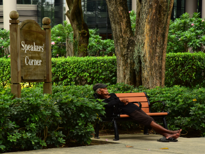 Ein Mann lehnt schlafend auf einer Bank im Park in Singapur. Neben ihm steht ein Schild, auf dem Speakers Corner steht.