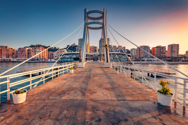 Sonnenuntergang an der Hafenpromenade von Durrës mit moderner Fußgängerbrücke und Blick auf Wohngebäude und das Meer. | Gebeco