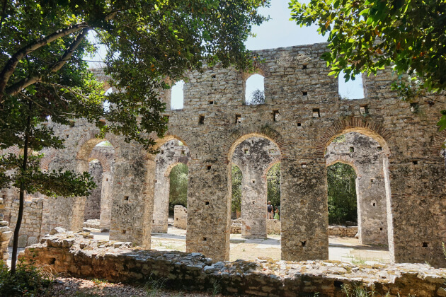 Antike Steinruinen der Basilika in Butrint, Albanien, mit Rundbögen und umgebenden Bäumen im Sonnenlicht. | Gebeco