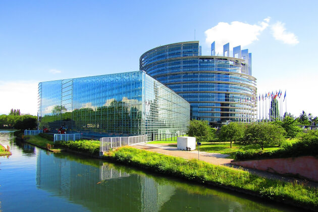 Europäisches Parlament in Straßburg mit moderner Glasfassade und EU-Flaggen am Flussufer bei Sonnenschein. | Gebeco