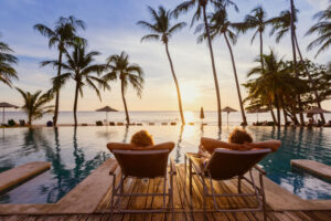 Paar entspannt auf Poolliegen mit Blick auf Infinity-Pool, Palmen und Sonnenuntergang am Meer