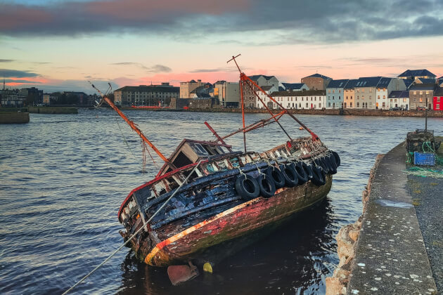 Verfallenes Holzschiff am Ufer von Galway vor bunten Häusern bei Sonnenuntergang. | Gebeco