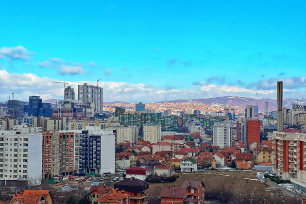 Stadtpanorama von Pristina mit modernen Hochhäusern, Wohnblöcken und Bergen im Hintergrund bei klarem Himmel. | Gebeco
