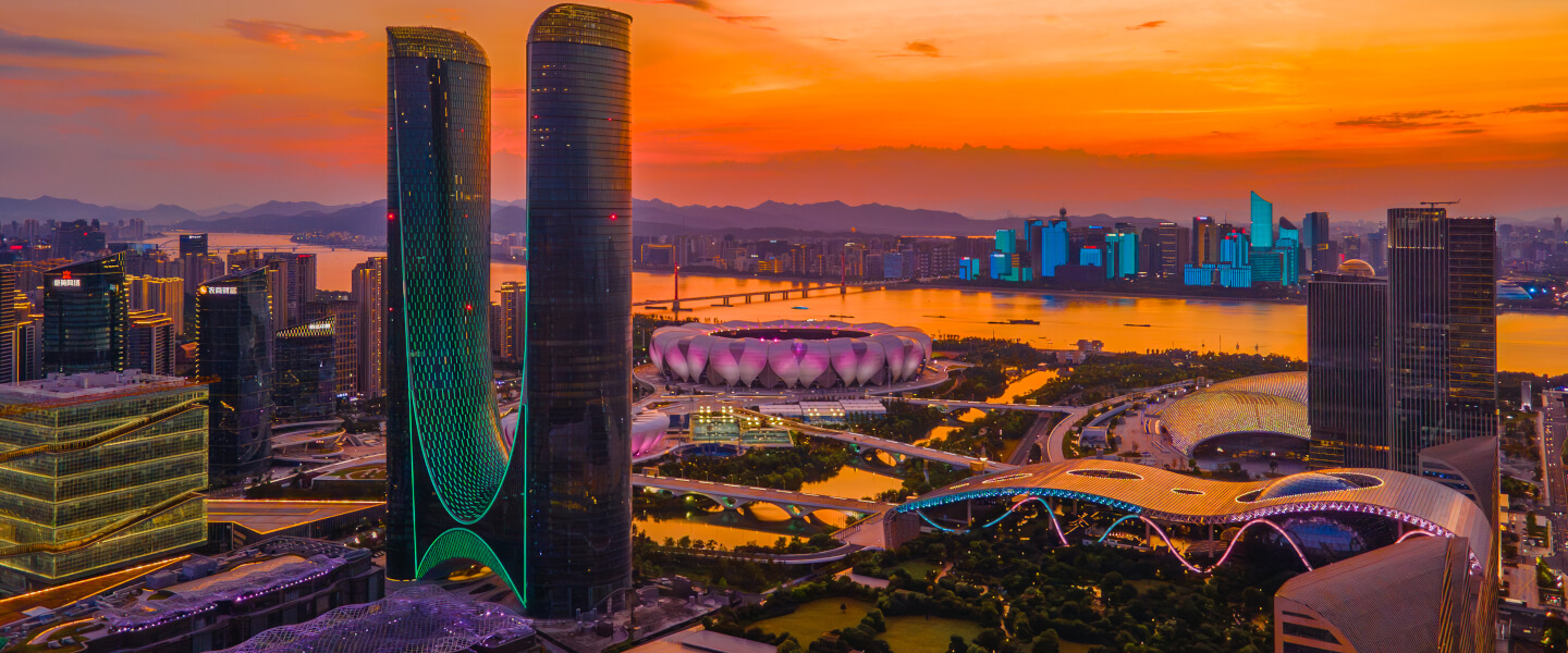 Moderne Skyline von Hangzhou mit markanten Hochhäusern, beleuchtetem Stadion und futuristischer Brücke bei Sonnenuntergang.