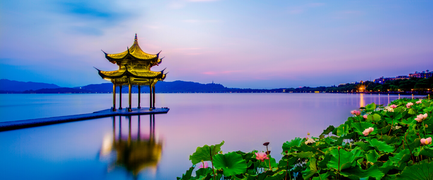 Goldener chinesischer Pavillon auf einem Steg im Westsee Hangzhou bei Sonnenuntergang, mit Spiegelung im Wasser und Lotusblumen im Vordergrund