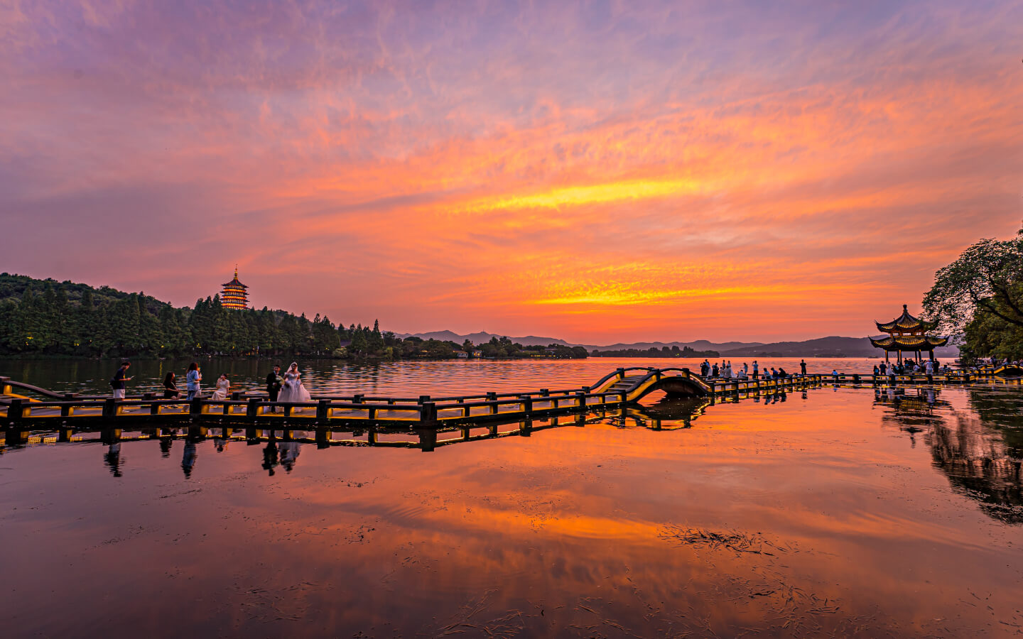 Sonnenuntergang am Westsee in Hangzhou mit langem Holzsteg, kleiner Bogenbrücke, goldfarbenem Pavillon und Leifeng-Pagode im Hintergrund. Menschen spazieren auf dem Steg und die Wasseroberfläche spiegelt den farbintensiven Himmel wider