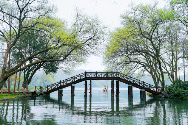 Holzbrücke am Westsee in Hangzhou im Frühling, umgeben von grünen Bäumen und ruhigem Wasser, chinesische Naturidylle
