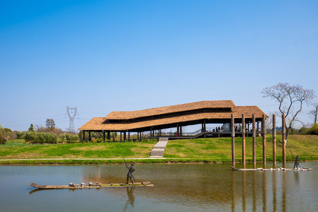 Modernes Pavillongebäude mit Strohdach an einem Fluss, davor Skulpturen auf Bambusflößen in ländlicher Landschaft unter blauem Himmel.
