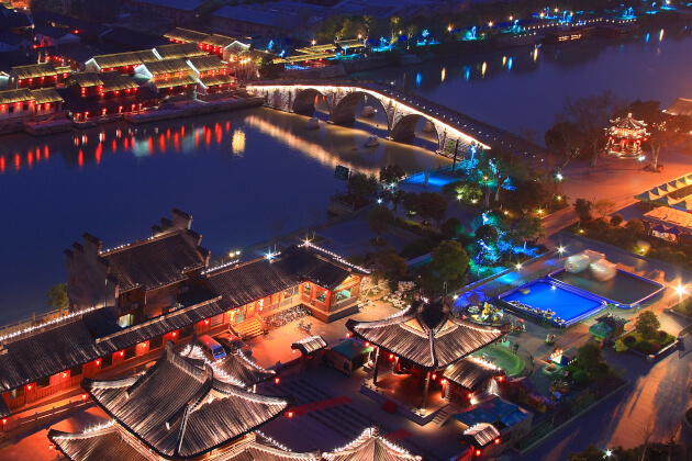 Traditionelle chinesische Altstadt bei Nacht mit beleuchteter Bogenbrücke über einen Fluss, historische Gebäude und bunte Lichtinstallationen.