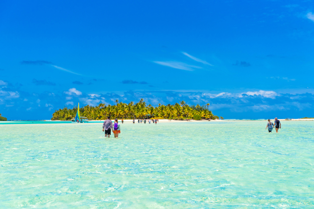 Menschen waten durch flaches, türkisfarbenes Wasser zu einer kleinen palmengesäumten Insel in Aitutaki auf den Cookinseln
