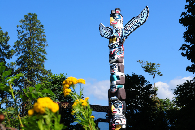Totempfahl im Stanley Park in Vancouver, umgeben von Bäumen und Blumen unter blauem Himmel