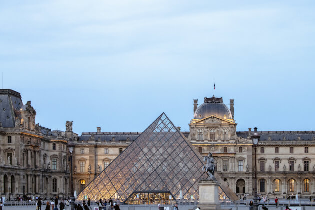Glaspyramide am Eingang des Louvre-Museums in Paris mit klassizistischer Fassade im Hintergrund bei Abenddämmerung. | Gebeco