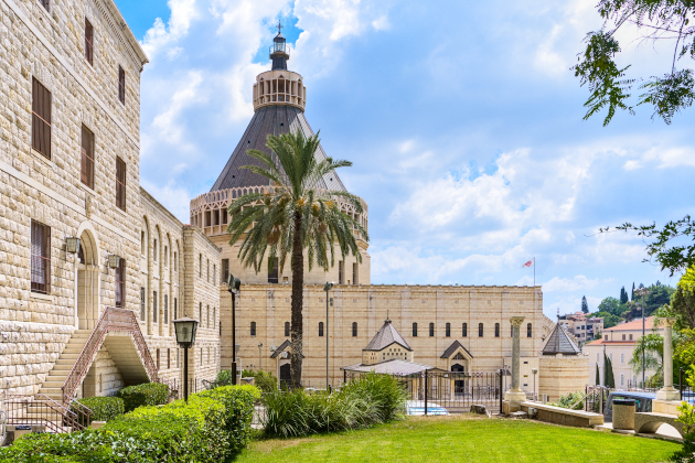 Basilika der Verkündigung in Nazareth mit großer Kuppel, hellem Sandsteinmauerwerk, Palmen und gepflegtem Vorplatz im Vordergrund.