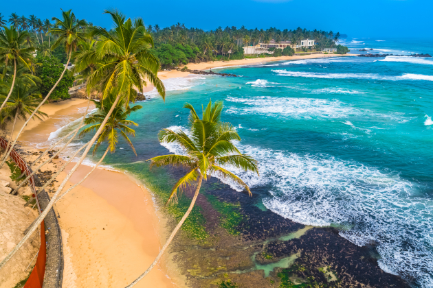 Strand von Unawatuna in Sri Lanka mit hohen Palmen, ruhiger Brandung und traditionellen Fischfang-Stelzen im Wasser. | Gebeco