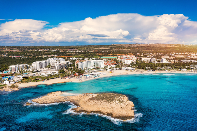 Luftaufnahme der Küste von Ayia Napa auf Zypern mit Sandstrand, türkisblauem Meer, Hotels und kleiner Felsinsel im Vordergrund.