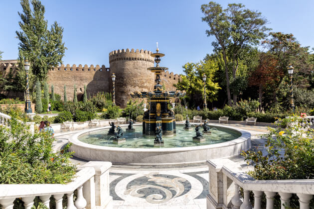 Historische Festungsmauer mit rundem Turm und Zinnen in Baku, Aserbaidschan, im Vordergrund gepflegter Park mit ornamentalem Springbrunnen und Gartenanlage.