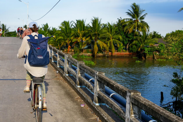 Radfahrerin mit Rucksack fährt über eine Betonbrücke entlang eines Flusskanals, gesäumt von Palmen und tropischem Ufergrün.