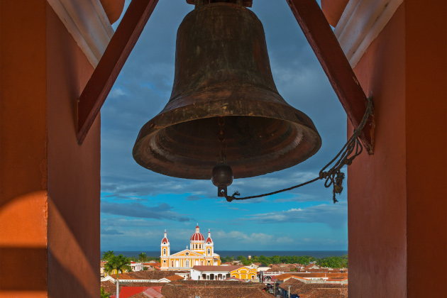 Blick durch eine Kirchenglocke auf die Kathedrale von Granada in Nicaragua mit Gebäuden aus der Kolonialzeit und blauem Himmel.