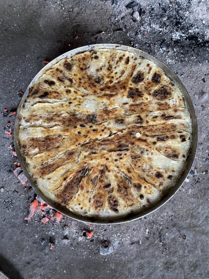 Rundes, goldbraun gebratenes Flija mit dunklen Grillflecken in Metallpfanne über glühender Kohle