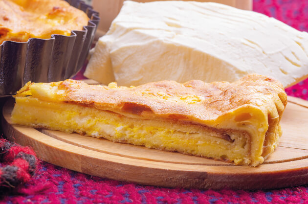 Stück herzhafte Tarte mit cremiger Käsefüllung auf Holzbrett, im Hintergrund Tarteform und großes Stück Weißkäse auf buntem Stoff