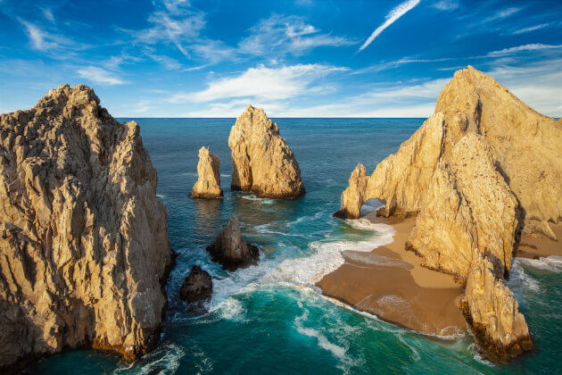 Panoramische Luftaufnahme markanter Felsformationen und Felsbogen an einer felsigen Küste mit türkisblauem Meer und kleinem Sandstrand unter blauem Himmel