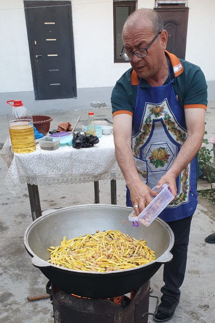 Reiseleiter bereitet traditionelles Plov in großem Kasan über offenem Feuer zu; Zutaten und Öl im Hintergrund.