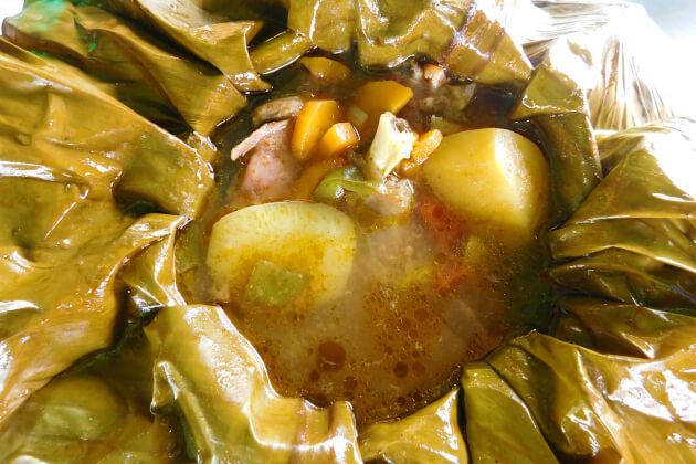 Nahaufnahme von Luwombo — einem traditionellen ugandischen Eintopf im Bananenblatt mit Kartoffelstücken, Maiskörnern, zarten Fleischstücken und aromatischer Brühe.