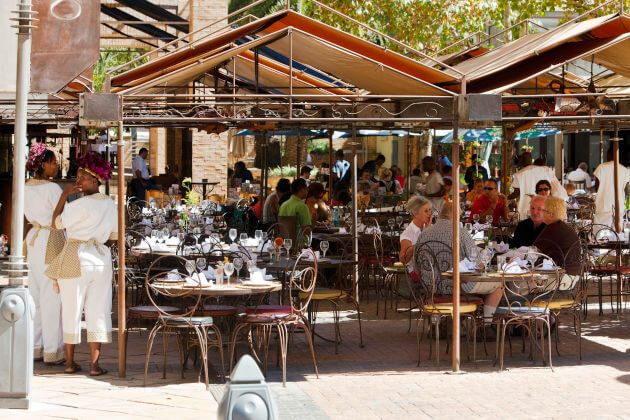 Belebtes Terrassenrestaurant am Melrose Square, Melrose (Johannesburg), Südafrika — gedeckte Tische, Gäste und Servicepersonal unter Markisen